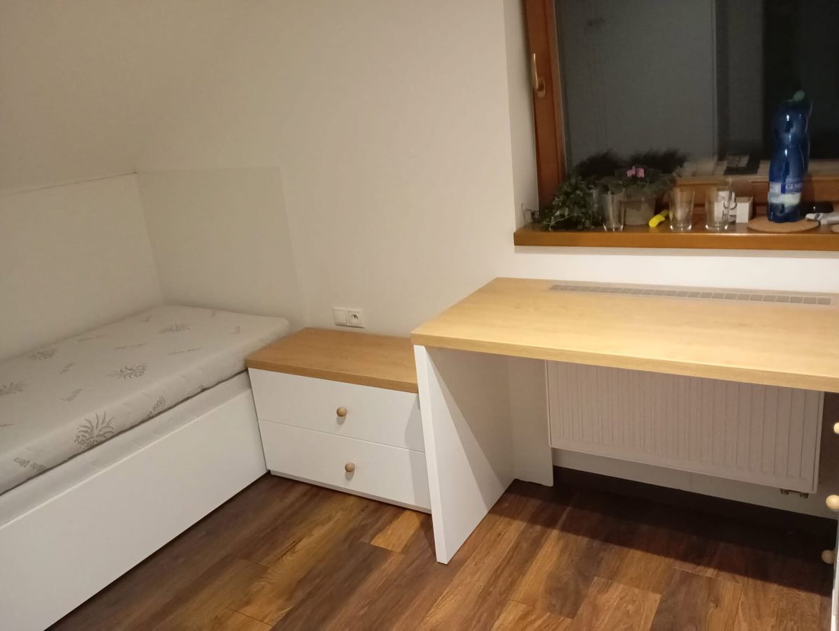 Lavica s matracom pod šikminou, vedľa nej zásuvka a písací stôl pod oknom